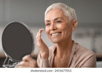 Anti Aging Skincare. Cheerful Senior Lady Applying Facial Moisturizer Preventing Wrinkles Looking At Round Mirror, Caring For Her Ageless Beauty At Home, Enjoying Self Pampering - Powered by Shutterstock