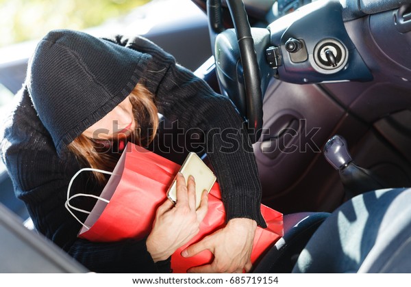 Anti theft system problem concept. Burglar thief man\
wearing black clothes breaking into car, stealing smartphone and\
red shopping bag