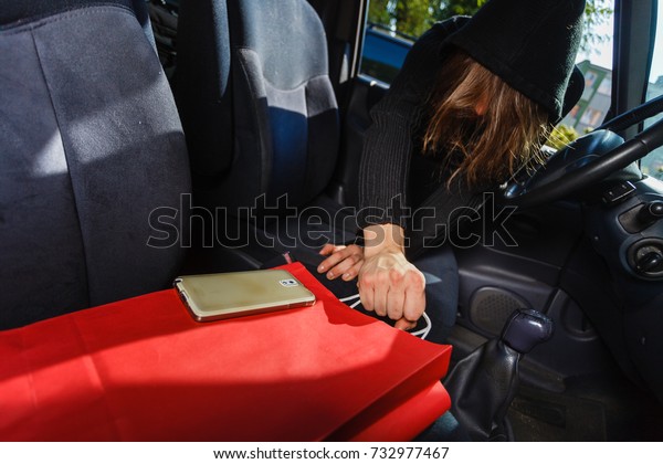 Anti theft system problem concept. Burglar thief man\
wearing black clothes breaking into car, stealing smartphone and\
red shopping bag