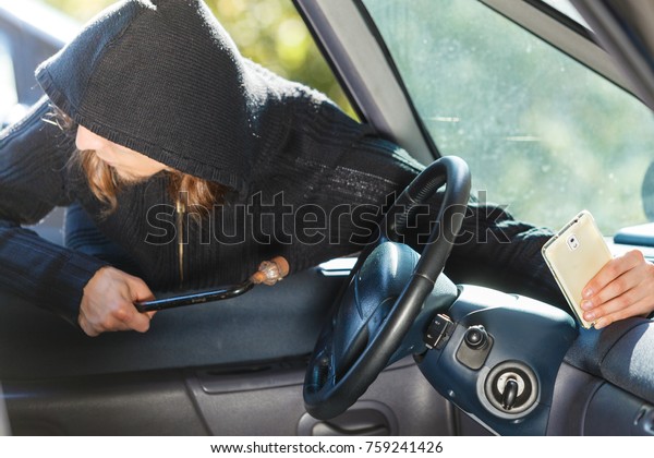 Anti\
theft system problem concept. Burglar thief man wearing black\
clothes breaking into car and stealing\
smartphone
