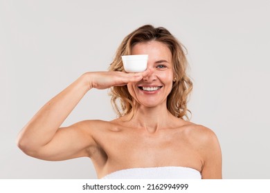 Anti-Age Cosmetic. Portrait Of Smiling Middle Aged Woman Wrapped In Towel Holding Jar With Moisturising Cream On Her Palm, Covering One Eye With New Skincare Product, Posing On Grey Background