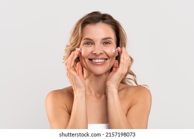 Antiage Treatment. Beautiful Caucasian Woman Applying Moisturizing Cream On Face, Attractive Millennial Female Nourishing Skin While Standing Wrapped In Towel Over Light Studio Background, Copy Space