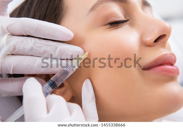 Anti-aging face treatment. Close up side on\
portrait of young smiling pretty woman relaxing on rejuvenation\
procedure of cheek bone zone by\
specialist