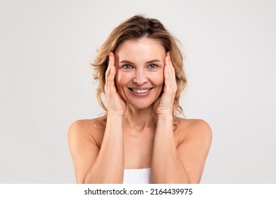 Anti-Aging Skincare. Beautiful Middle Aged Woman Touching Her Face And Smiling At Camera, Attractive Happy Mature Woman Standing Wrapped In Towel Over Light Grey Studio Background, Copy Space