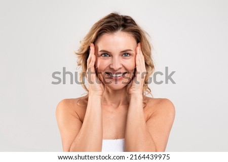 Anti-Aging Skincare. Beautiful Middle Aged Woman Touching Her Face And Smiling At Camera, Attractive Happy Mature Woman Standing Wrapped In Towel Over Light Grey Studio Background, Copy Space