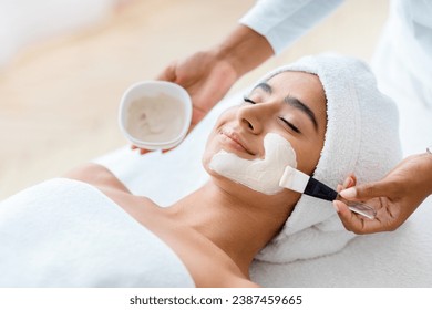 Anti-Aging Skincare. Cosmetologist applying clay mask on face of young indian woman, beautiful eastern lady with towel on head lying on table, enjoying beauty treatments in spa salon, closeup - Powered by Shutterstock