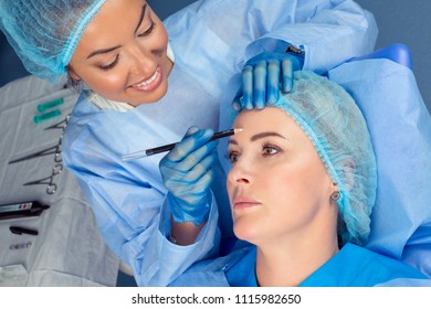 Anti-aging Treatment Face Lift. Beautiful Doctor Woman About To Draw Lift Line On Patient Forehead With Pencil For Anti-wrinkles Lifting Procedure. Two People In Surgery Clinic Room Patient Lying Down