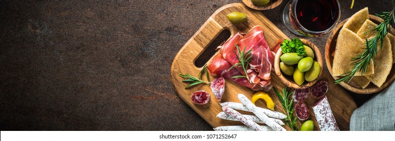 Antipasto - sliced meat, ham, salami, olives and glass wine on dark stone table. Top view long banner format.