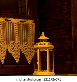 An Antique Candle Lantern Standing On A Wooden Windowsill. Wood And Brick Building Interior. Windows With A Wooden Frame, A Wooden Wall And A Window Sill. Visible Short Curtains With A Lace Structure.