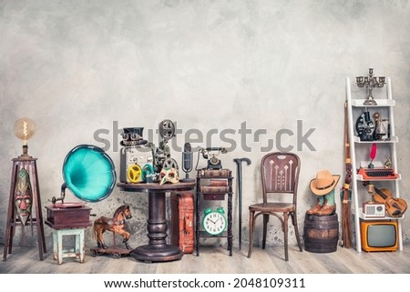 Antique gramophone, chair, retro microphone and recorder on table. Old telephone, typewriter, radio, TV on shelving front concrete wall background. Nostalgic still life. Vintage style filtered photo