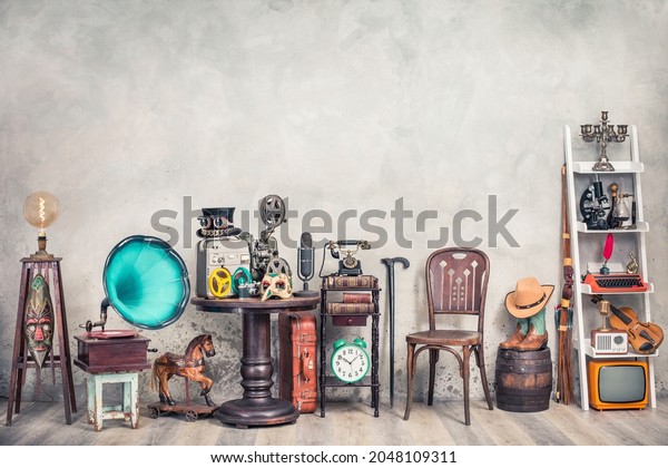 Antique gramophone, chair, retro microphone and\
recorder on table. Old telephone, typewriter, radio, TV on shelving\
front concrete wall background. Nostalgic still life. Vintage style\
filtered photo