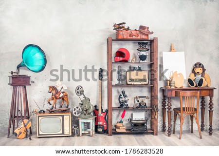 Antique media devices, writers tools, gramophone, film projector, old Teddy Bear toys and white canvas blank on easel, violin and guitar front concrete wall background. Vintage style filtered photo