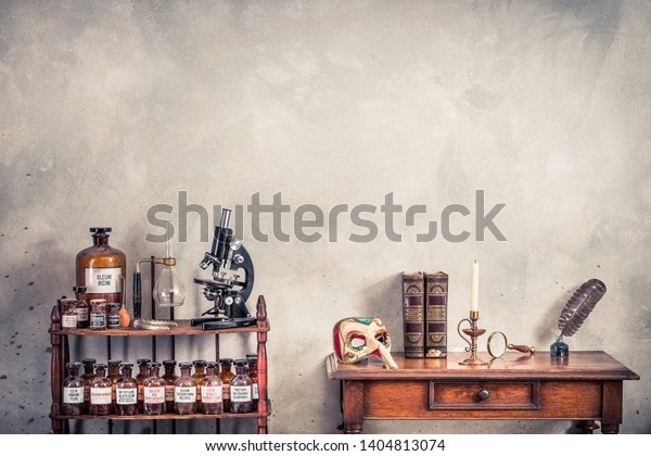 Antique microscope, old laboratory bottles on\
wooden shelving, carnival mask, quill ink pen, inkwell, books,\
magnifying glass, candle in vintage candlestick on oak table. Retro\
style filtered photo