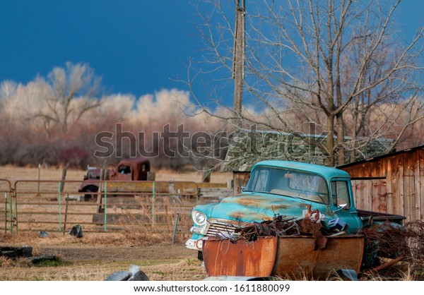 Antique\
rusty light blue pickup truck sits near a rusty junk pile near an\
aged wooden structure under the stormy dark\
sky