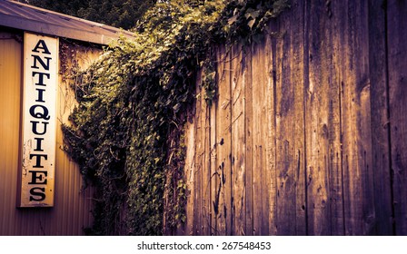 Antique Shop Sign Near Old Wooden Garden Wall Overgrown With A Ivy Shrub. Selective Focus On The Signboard. Toned Photo. Vignette.