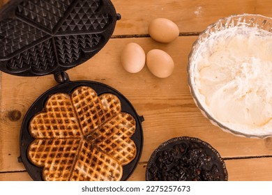 An antique waffle iron with a freshly baked waffle next to it are country eggs and a dish with freshly baked waffles and a bowl of blueberry jam and a bowl of whipped cream. High quality photo