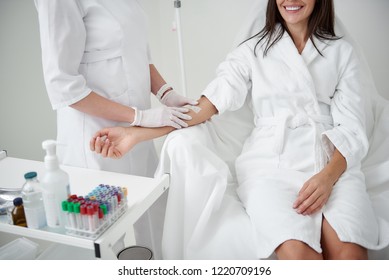 Anti-stress Procedure. Cropped Portrait Of Female Doctor In Sterile Gloves Preparing Lady Hand For IV Infusion. Brunette Girl In White Bathrobe Smiling