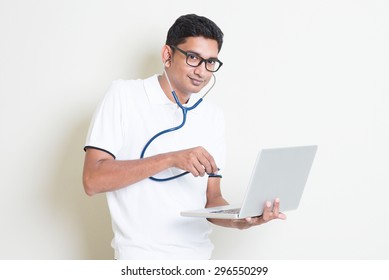 Antivirus Concept. Indian Computer Engineer Diagnose Virus Infected Pc With Stethoscope. Asian Man Standing On Plain Background With Shadow And Copy Space. 