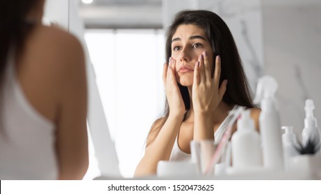 Anti-Wrinkle Skincare. Sad Young Woman Touching Face Looking At Her Skin In Mirror At Bathroom. Selective Focus, Panorama