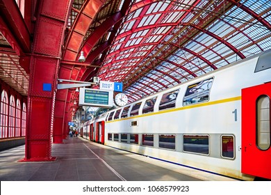 Antwerp, Belgium. Central indoor railway station. Platform made of red metal constructions with clock and panel with departure or arrival schedule. Modern double decker high-speed train.