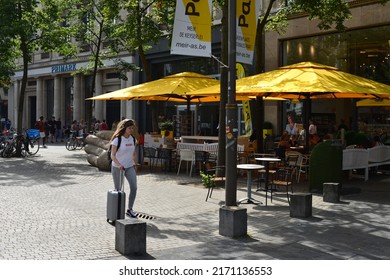 Antwerp city, Province Antwerp, Belgium- June 18, 2022: caucasian girl age 18 with luggage walking on Meir in front of bakery shop Panos with yellow parasols, outdoor tables and chairs on a sunny day
