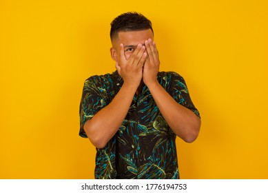 Anxiety - a conceptual image of a beautiful young caucasian man covering his face with his hands and peering out with one eye between her fingers standing indoors. Scared from something or someone.