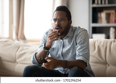 Anxious African American Young Man In Glasses Sit Relax On Couch In Living Room Watch TV, Biracial Millennial Male Rest On Sofa At Home Feel Worried About Bad Television News Or Program