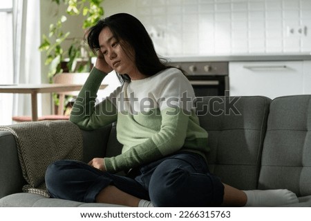 Anxious Asian millennial girl sitting on sofa feeling lonely, suffering from monophobia. Sad depressed young Korean woman sits alone at home looking aside, having problems dealing with panic attack