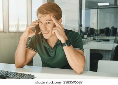 Anxious male employee sitting at table and touching forehead while speaking on phone in modern workspace