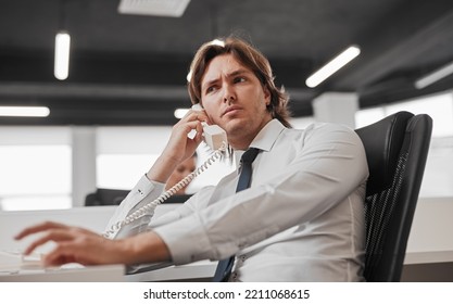 Anxious Man In Formal Wear Frowning And Looking Away While Sitting At Desk And Talking On Telephone During Work In Office