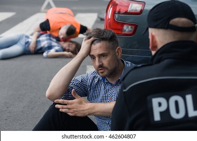 Anxious Man Sitting In Front Of The Police Officer With The Victim Of A Crash At The Background