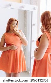 Anxious Oversized Young Woman Wearing Dress Stands In Front Of Mirror Looking Desperately At Her Reflection Holding Hand On Stomach And Eating Sweet Donut.