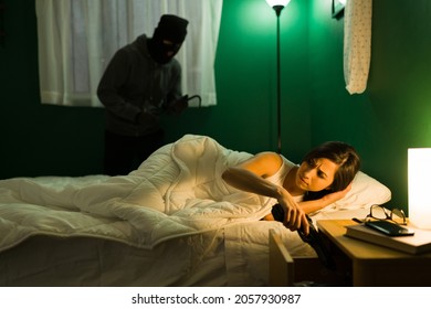 Anxious And Scared Woman In Bed Taking Out Her Gun From A Night Stand To Defend Herself From A Man Burglar At Her Home