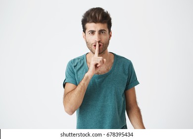 Anxious serious man wearing casual t-shirt holding index finger at his lips, asking to keep silent and not to make noize. Human facial expressions.