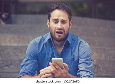 Anxious Upset Young Scared Man Looking At Phone Seeing Bad News Or Text Message Sitting On Stairs Outside Corporate Building. Human Emotion, Reaction, Expression