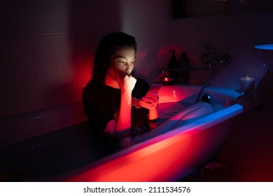 Anxious woman sit in clothes in bath of cold water reading text messages from ex-boyfriend, deleting photos with lover after breakup or betrayal. Angry and jealous girl with smartphone in bathroom