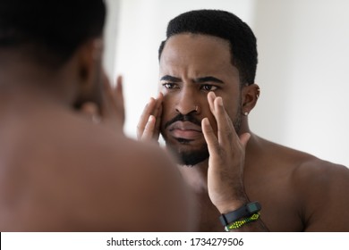 Anxious young african American man look in mirror in bathroom concerned about face wrinkles, frustrated unhappy biracial male worried about acne, unhealthy facial skin, skincare concept