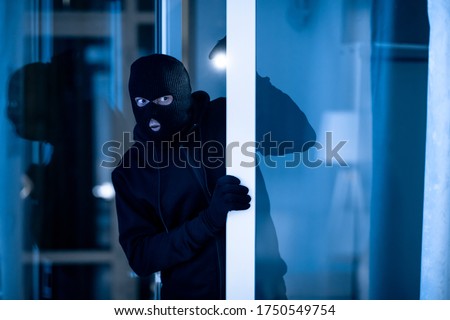 Is Anybody Home. Cautious masked thief looking and peeping into office through window or glass door with torch flashlight