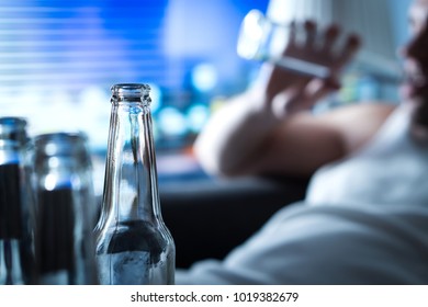 Anynomous Alcoholic Taking Sip Of Beer. Home Full Of Empty Bottles. Alcoholism And Drinking Problem Concept. Man Sipping Alcohol Late At Night. Drunken Sad Guy Sitting On Sofa.