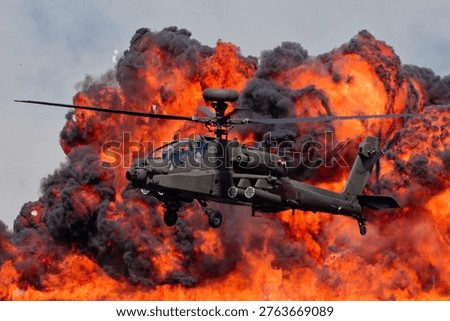 An Apache attack helicopter in flight against a backdrop of a large, controlled explosion, featuring vibrant orange flames and thick black smoke.