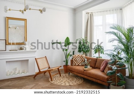 Apartment with bright living room in boho style. Interior design with brown couch, new armchair, decorative fireplace with candles, green house plants in clay pots and rug on the wooden floor