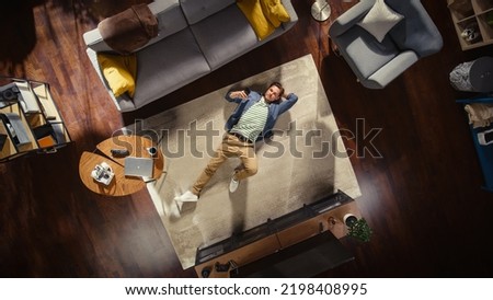 Apartment: Handsome Man Lying on a Living Room Floor, Using Mobile Phone. Guy Relaxes on the Carpet and Looking Up at Camera. Freelance, Remote Work, Online Shopping, Social Media Browsing. Top View.