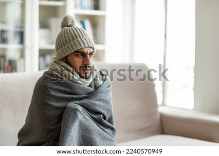 Apartment heating problem concept. Freezing angry middle eastern young guy sitting on couch in living room, wearing knitted hat, covered in warm blanket, warming up, looking at copy space