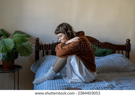 Apathetical unemotional introverted teenager sitting on bed embracing knees alone at home. Friendlessness, solitude, depression, absence of motivation, loss of life interest, self-destruction concept
