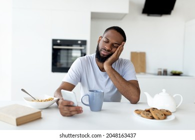 Apathy. Portrait of tired sleepy black man using smartphone while having breakfast in kitchen at home, bored African American male sitting at table, reading text messages on his cell phone eating food