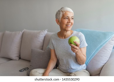An aple a day keeps doctor away. Close up of a cheerful elderly woman eating an apple while smiling in the livingroom. Healthy mid age woman holding apple closeup
