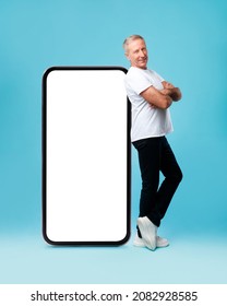 App Ad. Full Body Length Of Smiling Confident Adult Man Standing With Folded Arms Near Giant Cell Phone, Empty White Screen Template, Blue Studio. Mock Up For Website, Free Copy Space For Advertising