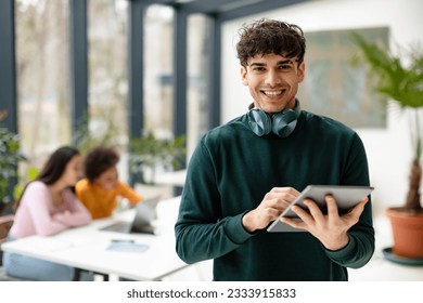 App for knowledge. Cheerful european guy students using tablet for video lesson, studying with friends in classroom, ladies sitting at table on background - Powered by Shutterstock