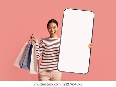 App For Online Store. Excited Happy Asian Woman Showing Big Giant Cell Phone With Blank White Screen In Hand, Lady Holding Shopping Bags, Isolated On Pink Studio Background, Mock Up, Free Copy Space