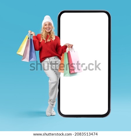 App For Online Store. Full Body Length Of Happy Woman In Winter Hat Standing Near Big Smartphone With Blank White Screen Holding Shopping Bags, Isolated Blue Background, Mock Up Image, Free Copy Space